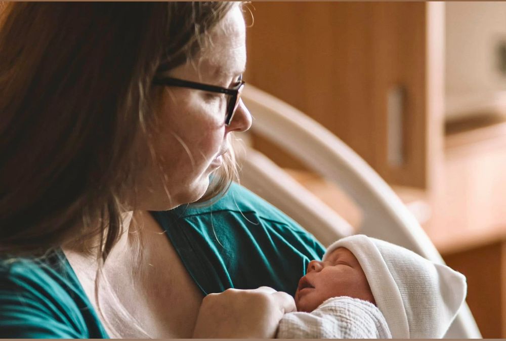 New mother with glasses tenderly holding her swaddled newborn baby in a hospital room.