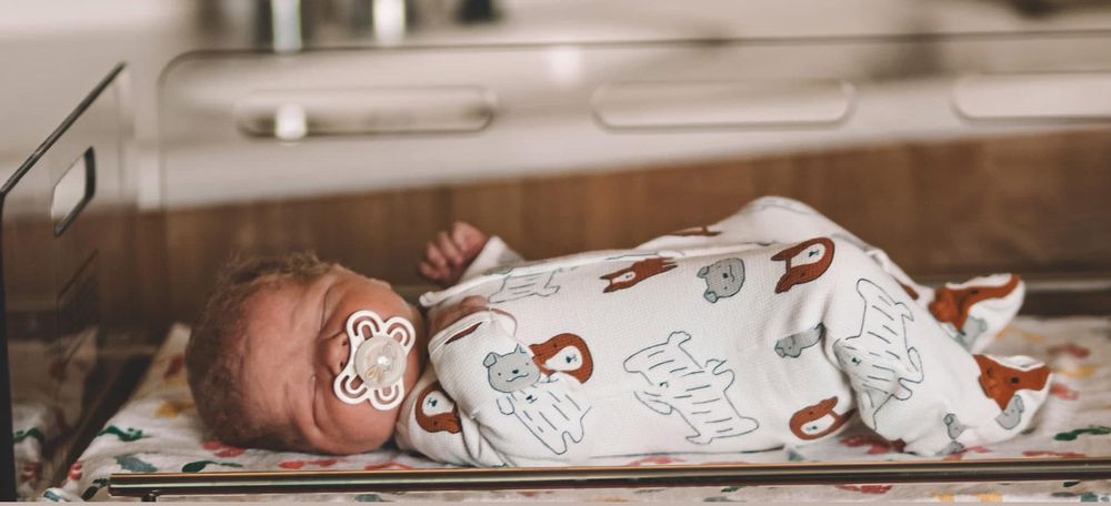 Newborn baby with a floral pacifier lying in a hospital bassinet wearing an animal print sleeper.