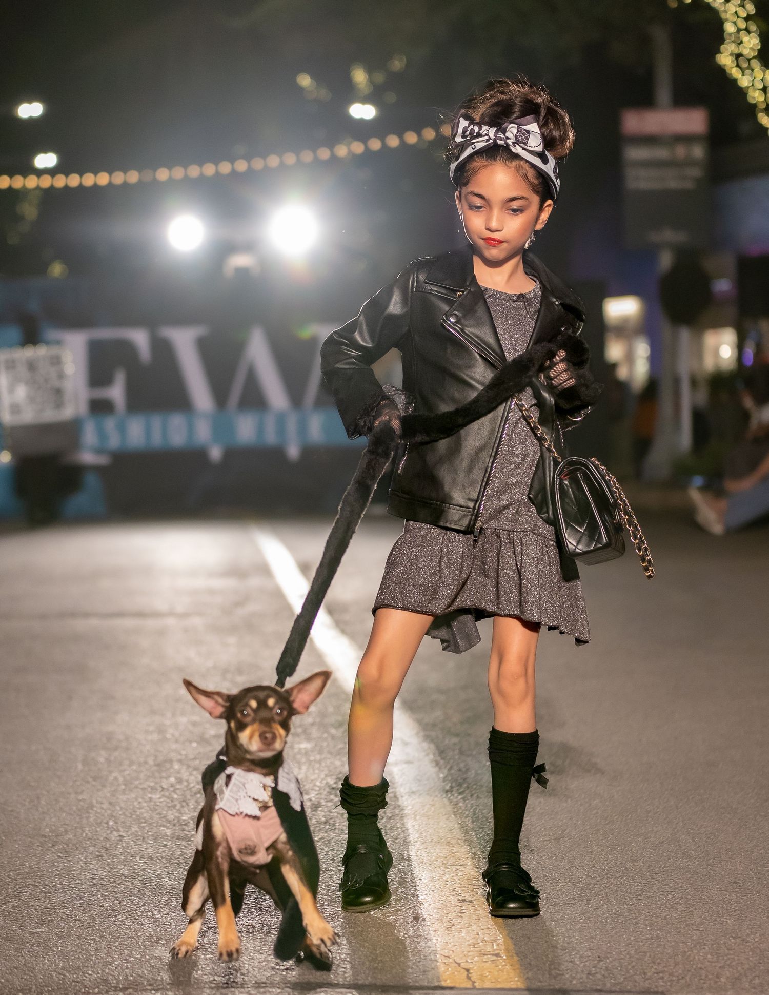 A young model with decorated hair walks a small well-dressed dog down a nighttime fashion runway.
