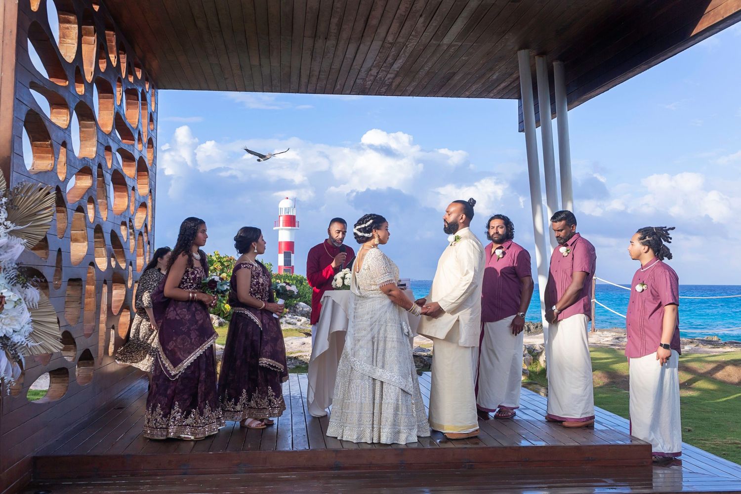 Bride and groom hold hands during a vibrant South Asian wedding ceremony at Hyatt Ziva Cancun, with the ocean and lighthouse.