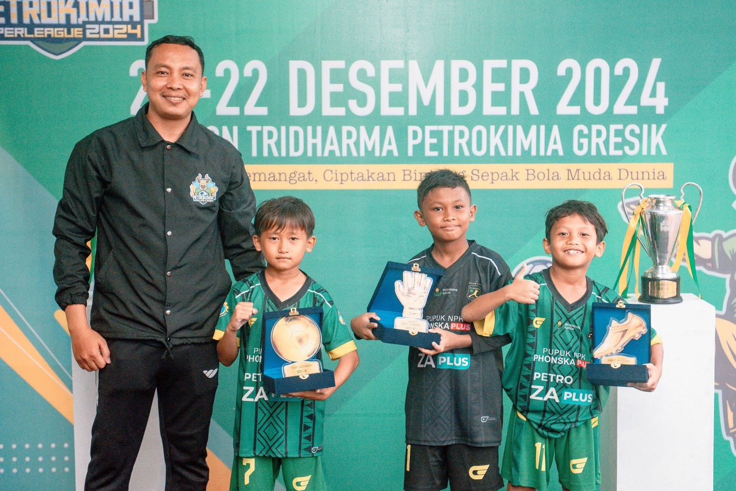 Young soccer players in green jerseys proudly display their trophies at a Petrokimia Gresik sports event.