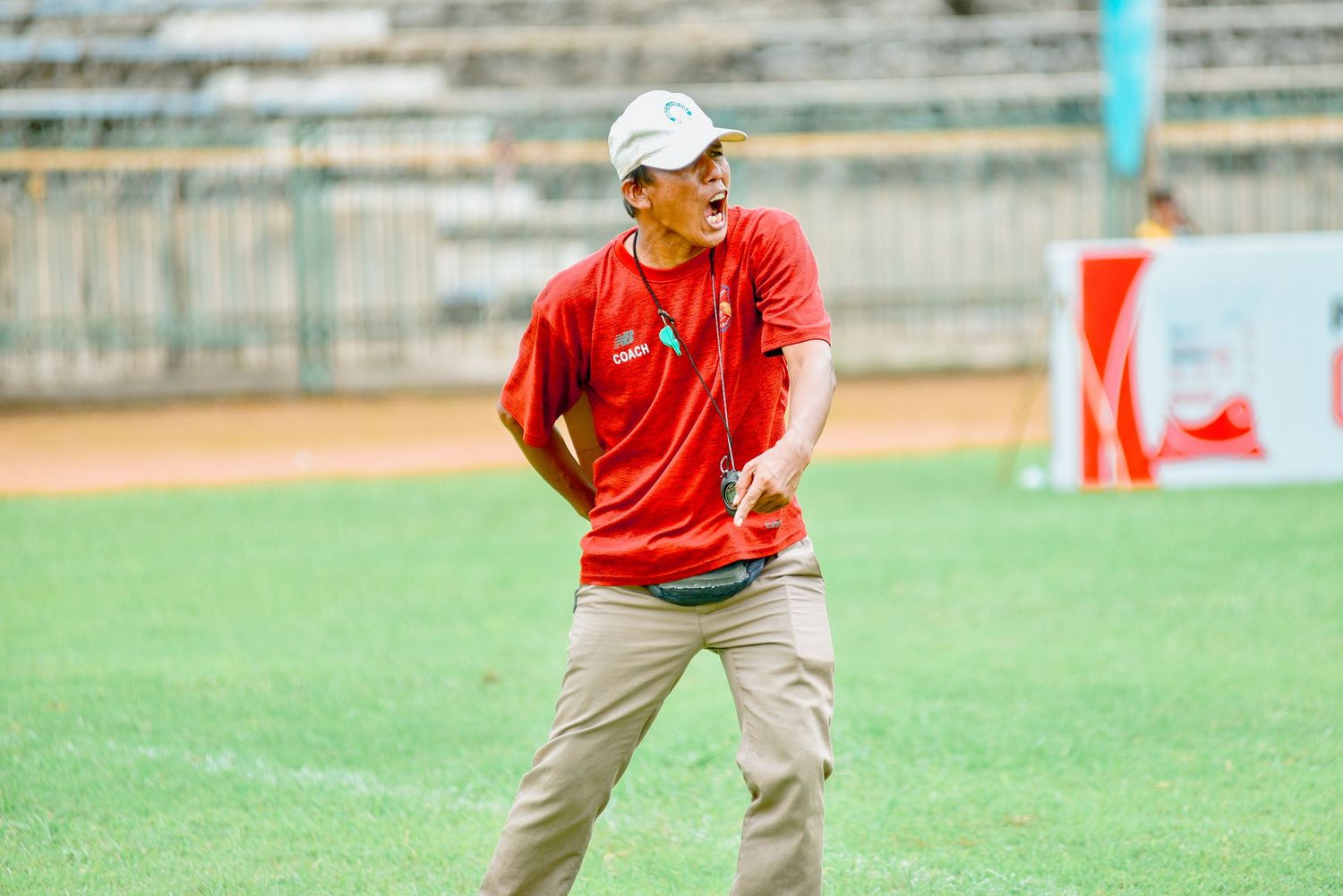 A football coach in red shirt and khaki pants gestures animatedly during a game on the sidelines.
