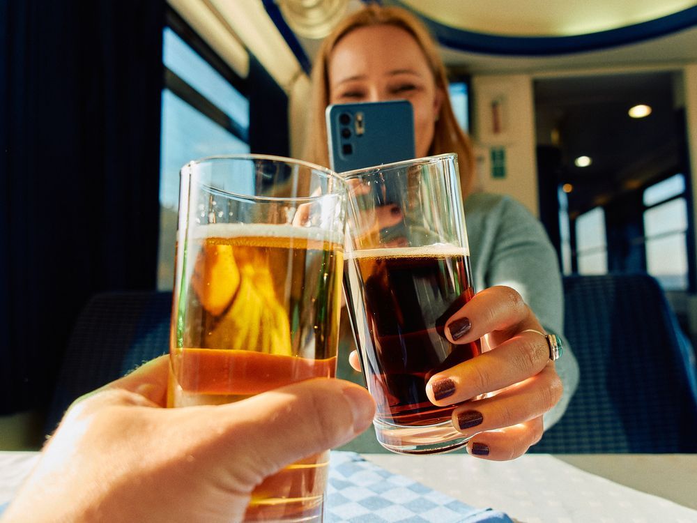 Two people clink glasses filled with different colored beers together while sitting on a train.