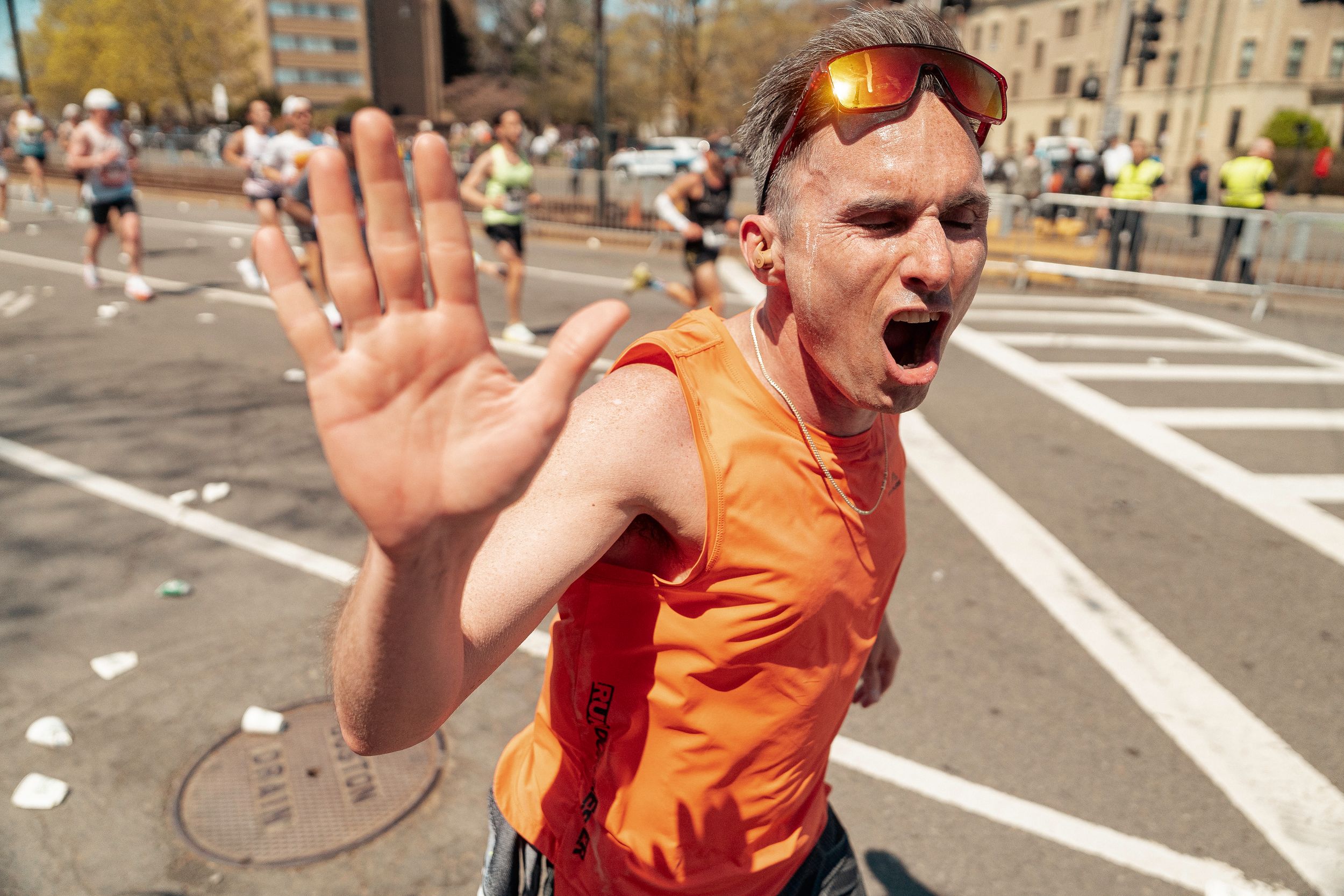 A marathon participant in an orange tank top gestures enthusiastically during a race on a city street.