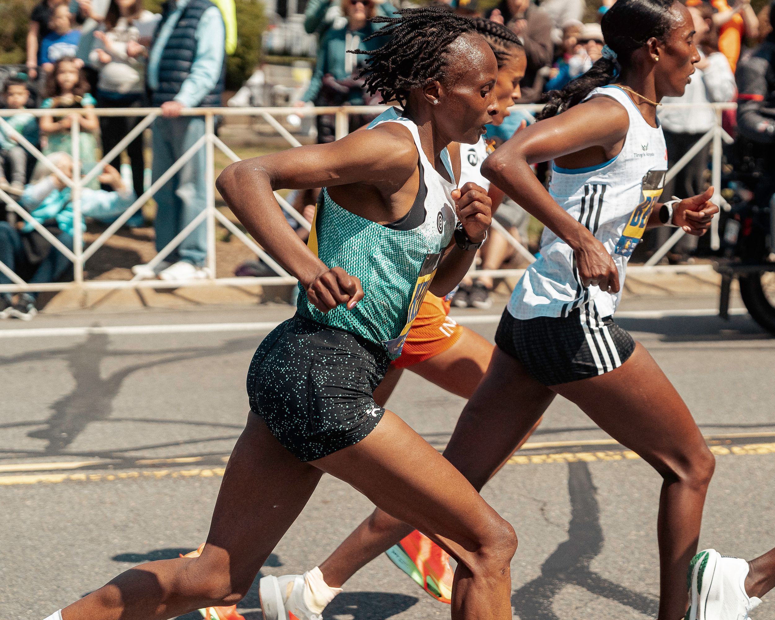 Two runners compete in a street race wearing athletic gear as spectators watch from behind barriers.