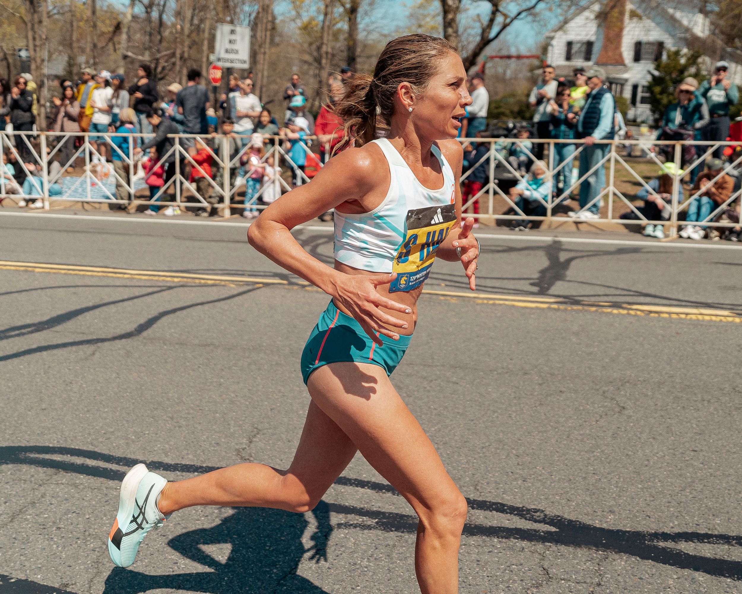 Runner in a light blue singlet and shorts competes on a sunny day as spectators watch from behind barriers.