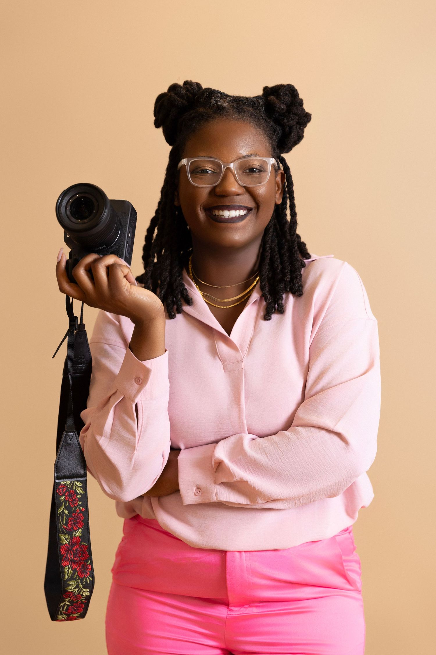 Creative photographer wearing a pink sweater and glasses holds a professional camera against beige backdrop.
