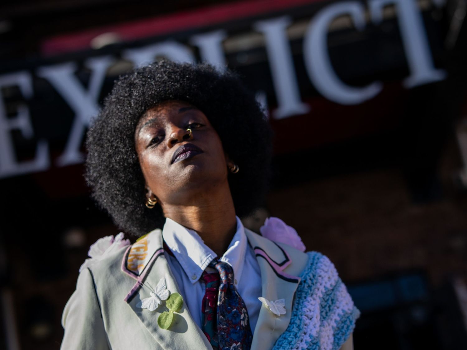 Person with afro hairstyle posing confidently outdoors near a sign, wearing layered eclectic clothing with colorful accessories.