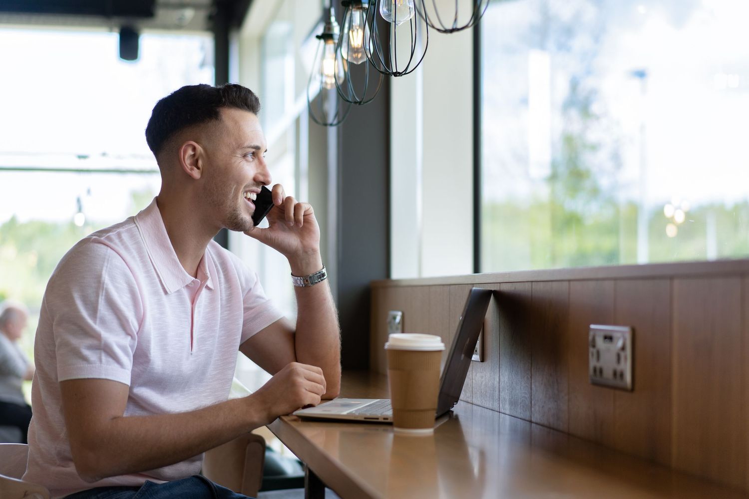 Male entrepreneur in polo shirt sits at cafe table with laptop and coffee while talking on phone near window. Personal branding photoshoot