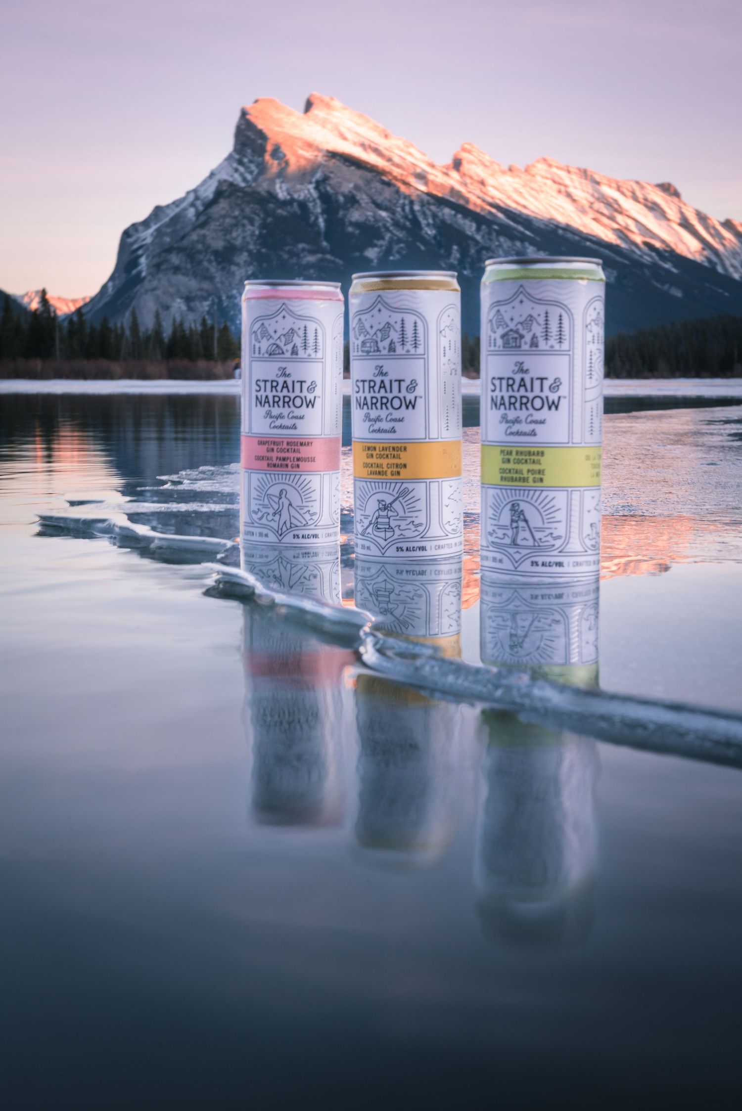 Three hard seltzer cans sit on frozen lake ice with snow-capped Mount Rundle reflecting in Banff National Park at sunset.