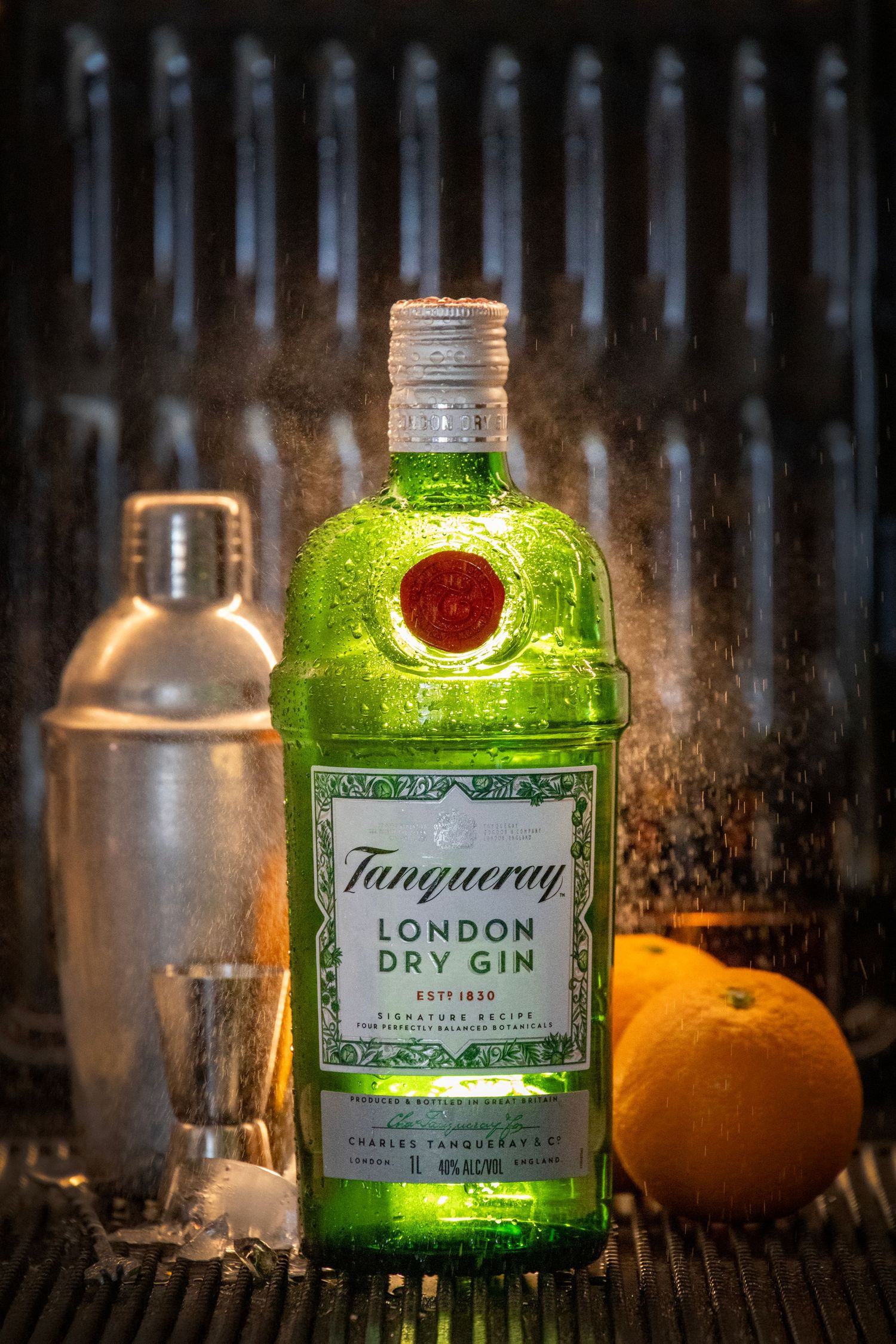 Tanqueray London Dry Gin bottle with cocktail shaker, shot glass, and oranges on a bar surface with moody backlighting.