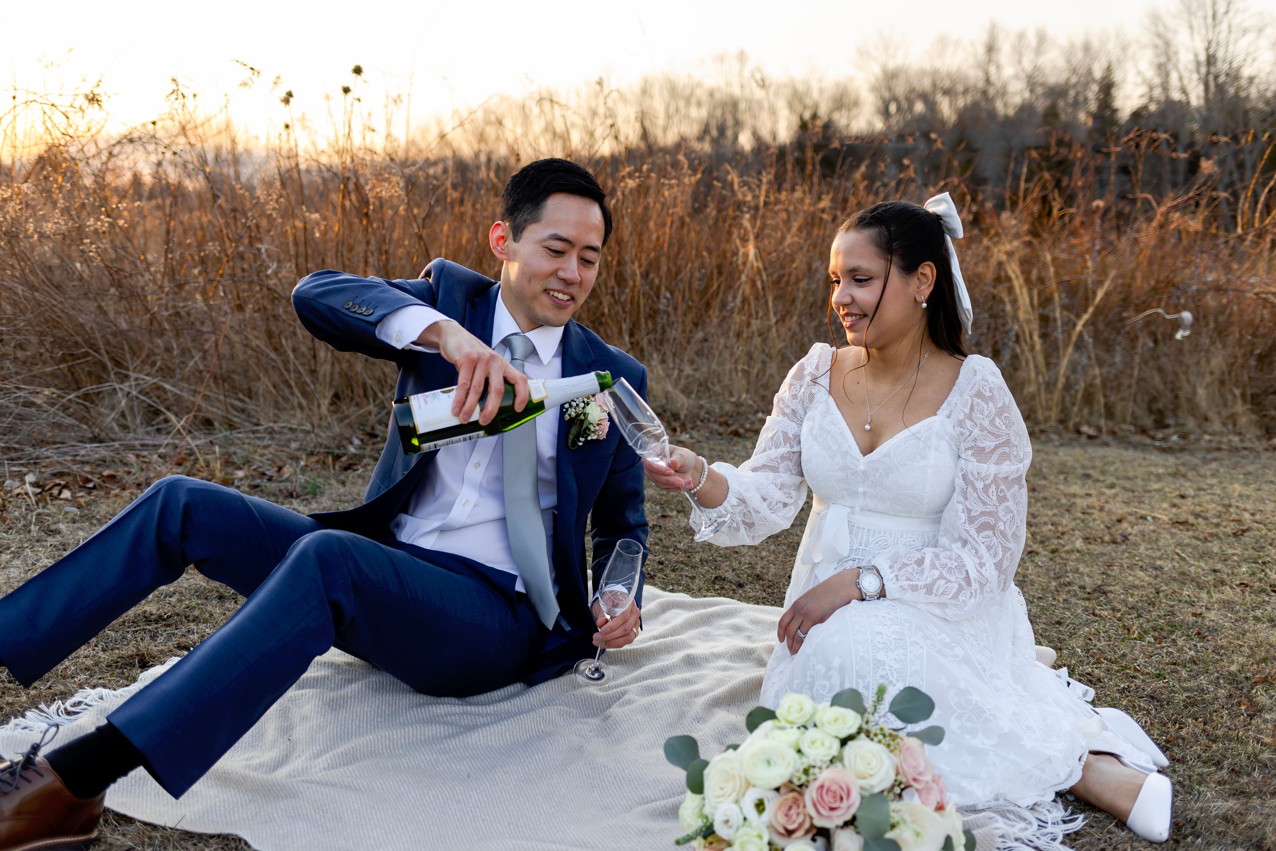 A couple celebrates their wedding day with champagne while sitting on a picnic blanket in a rustic outdoor setting at sunset.