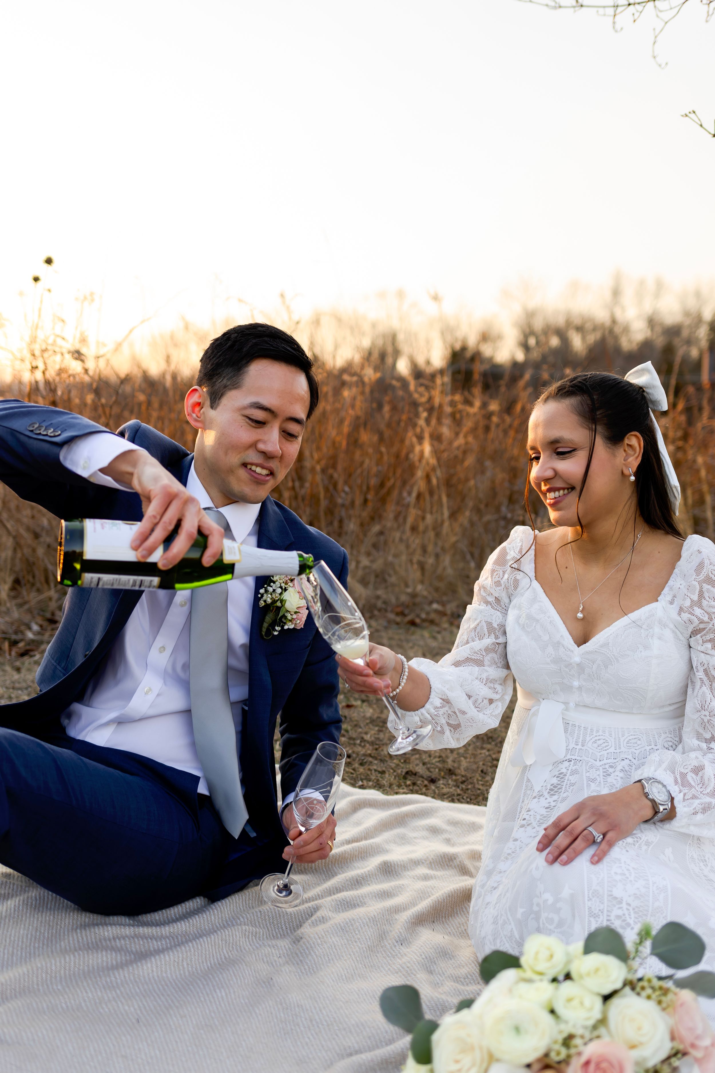 A newlywed couple shares a champagne moment on a blanket outdoors with floral bouquet during golden hour.