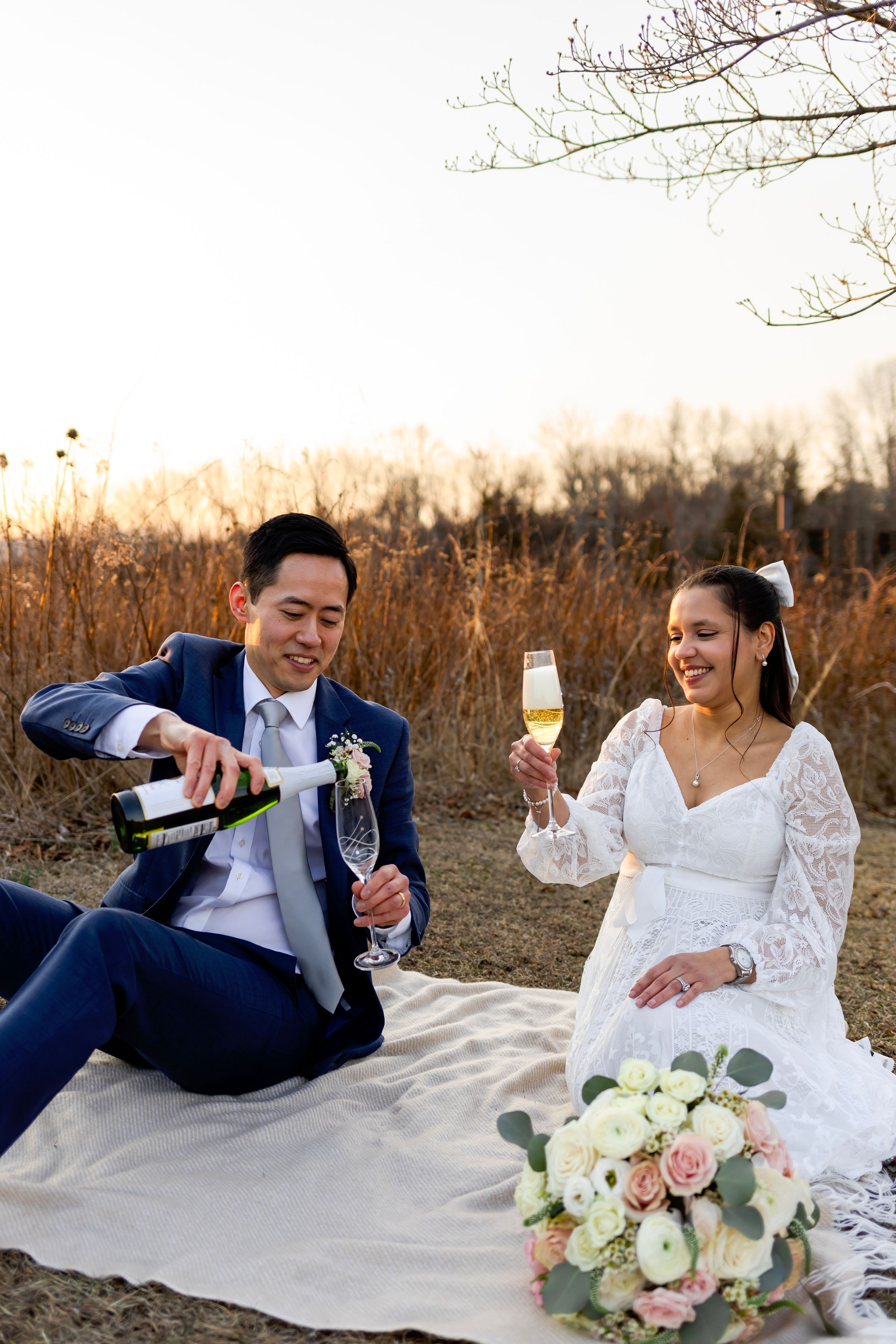 Newlyweds share champagne while sitting on picnic blanket with bridal bouquet of white and pink roses at sunset.