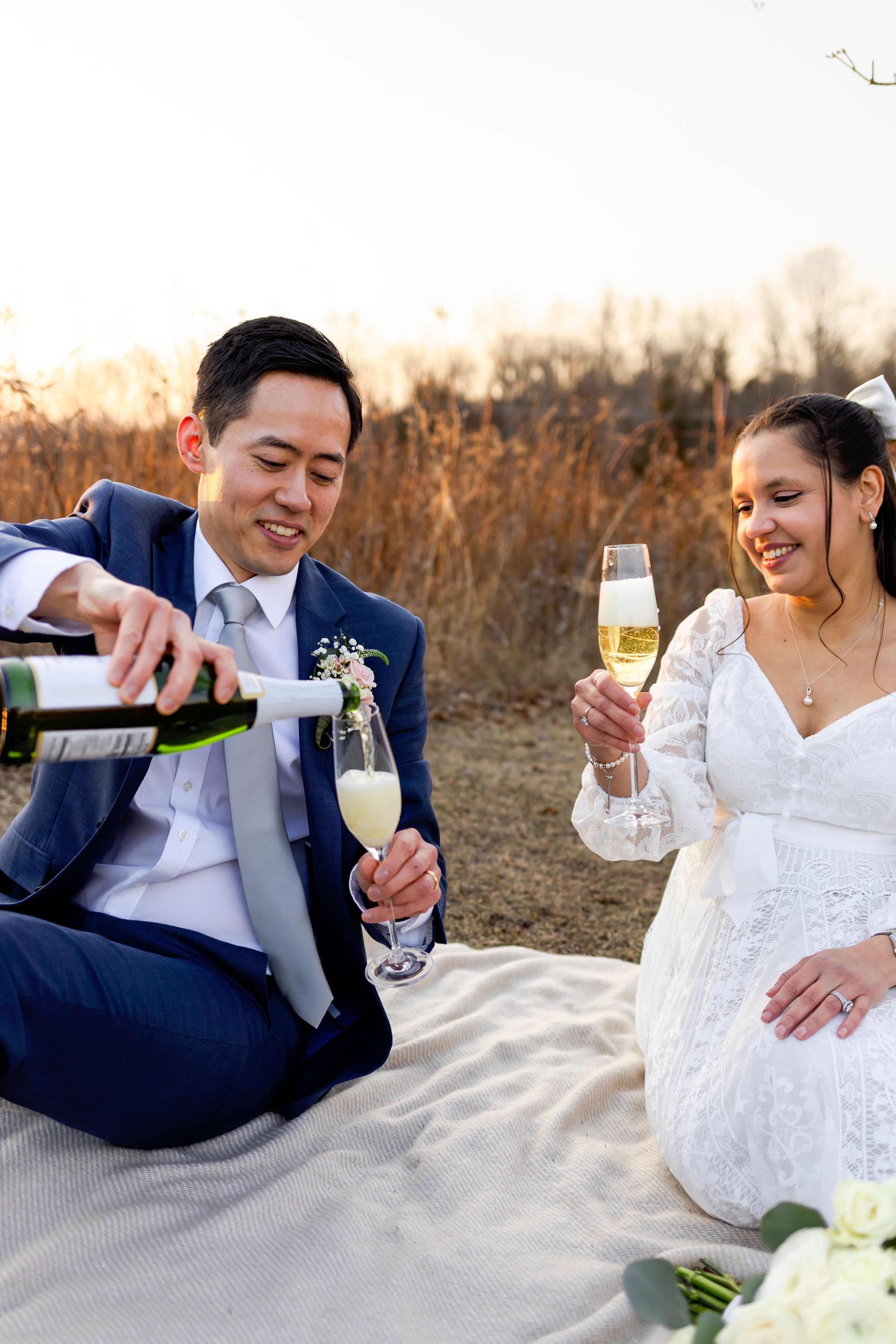 A newlywed couple pours champagne while sitting on a blanket outdoors during their wedding celebration.
