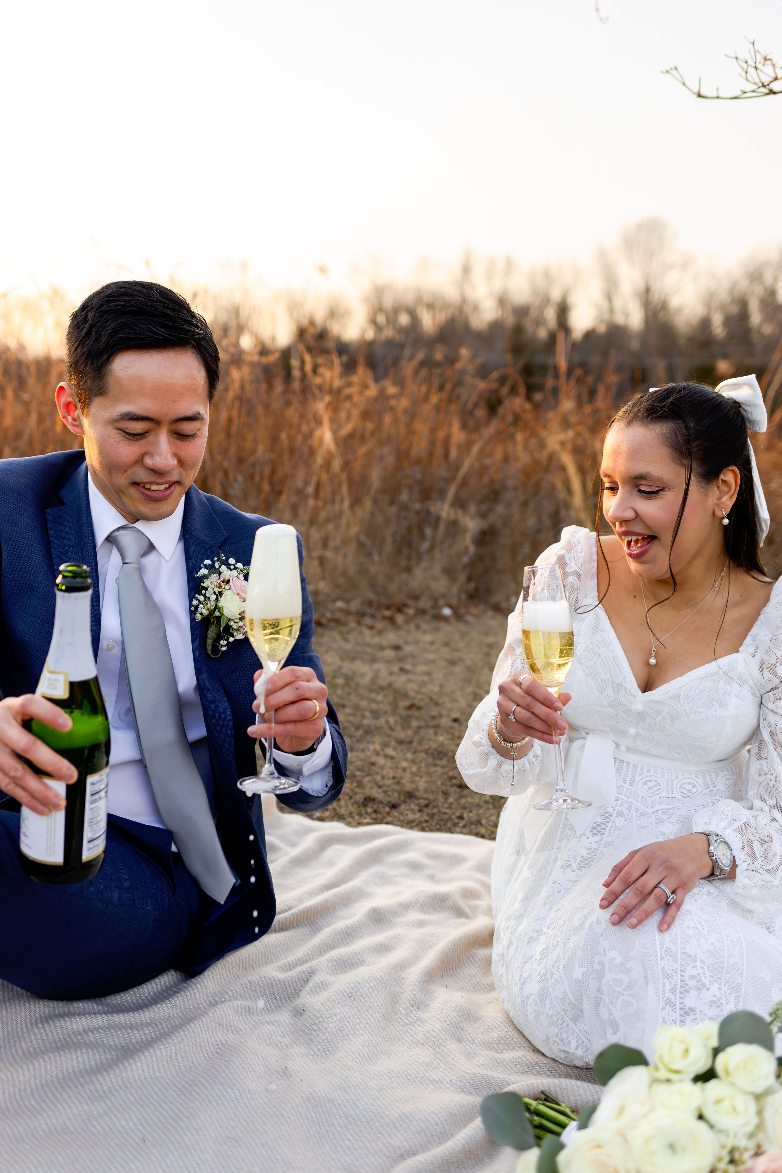 A couple toasts with champagne glasses while sitting on a picnic blanket with flowers at sunset.