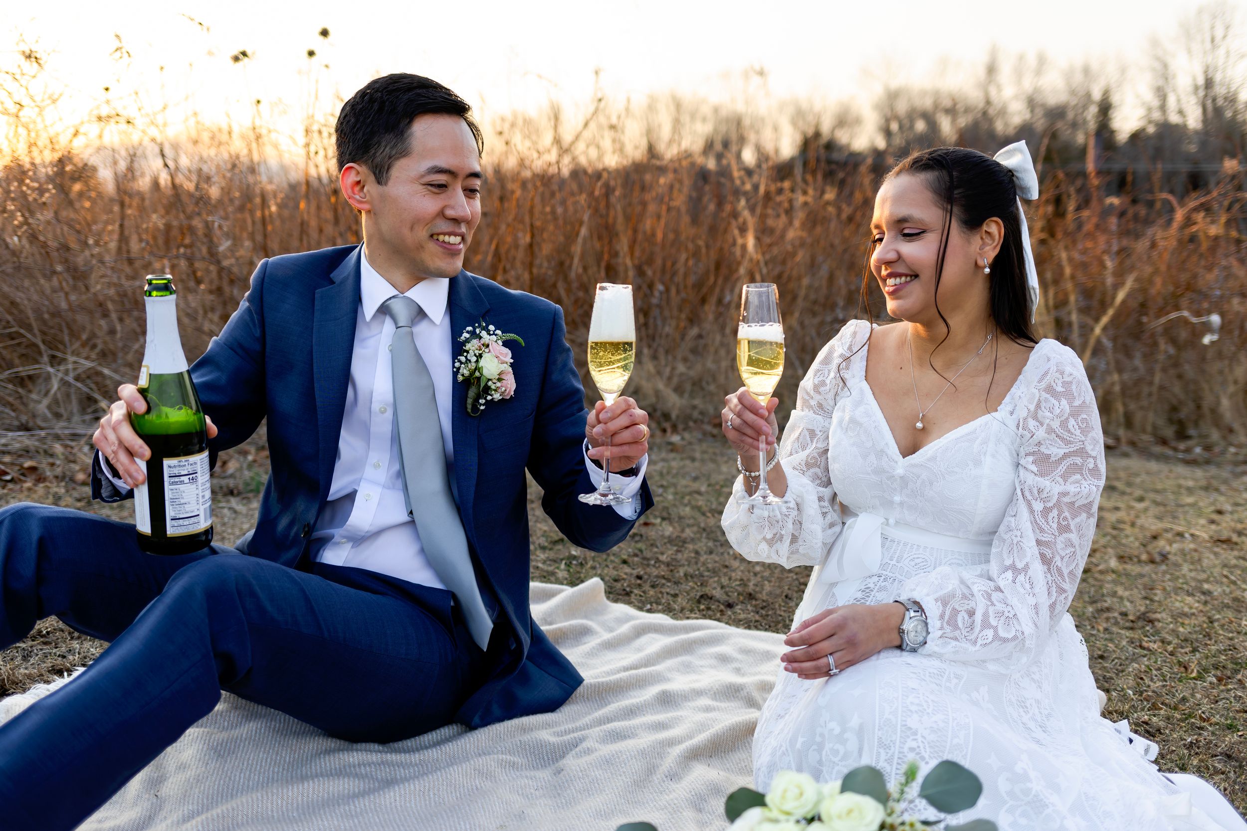 Newlywed couple toasts with champagne during outdoor sunset picnic celebration with bottles and flowers.