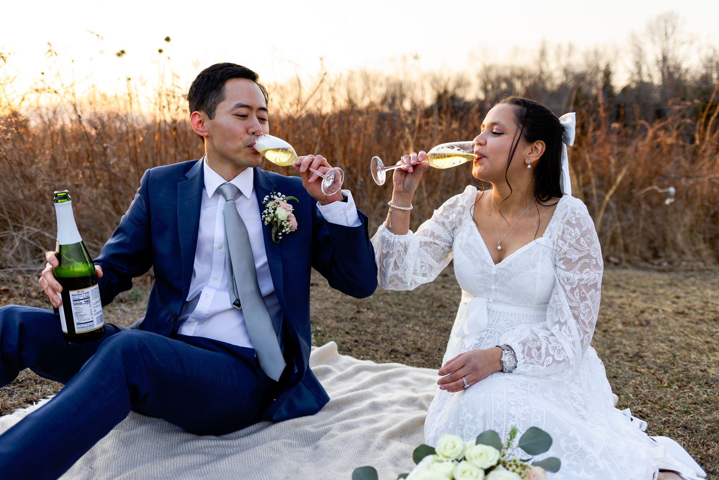 A couple in wedding attire sits on a blanket in a golden field sharing drinks from champagne glasses at sunset.
