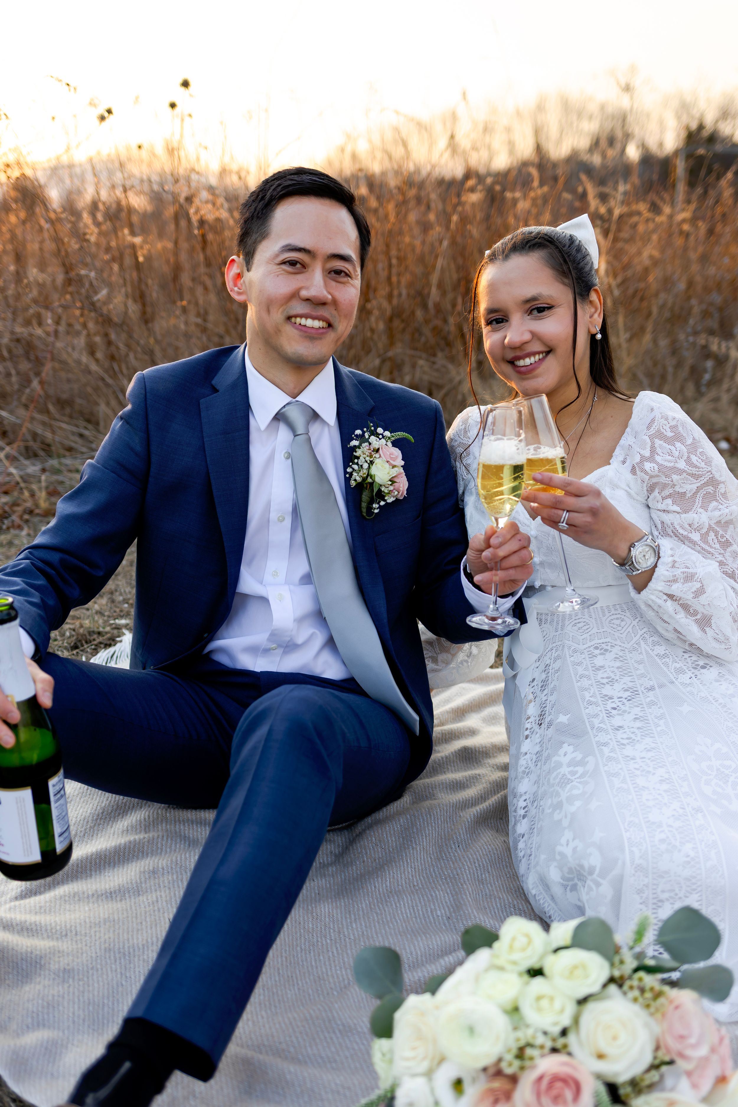 A couple in wedding attire having champagne outdoors with white roses and floral bouquet at sunset.