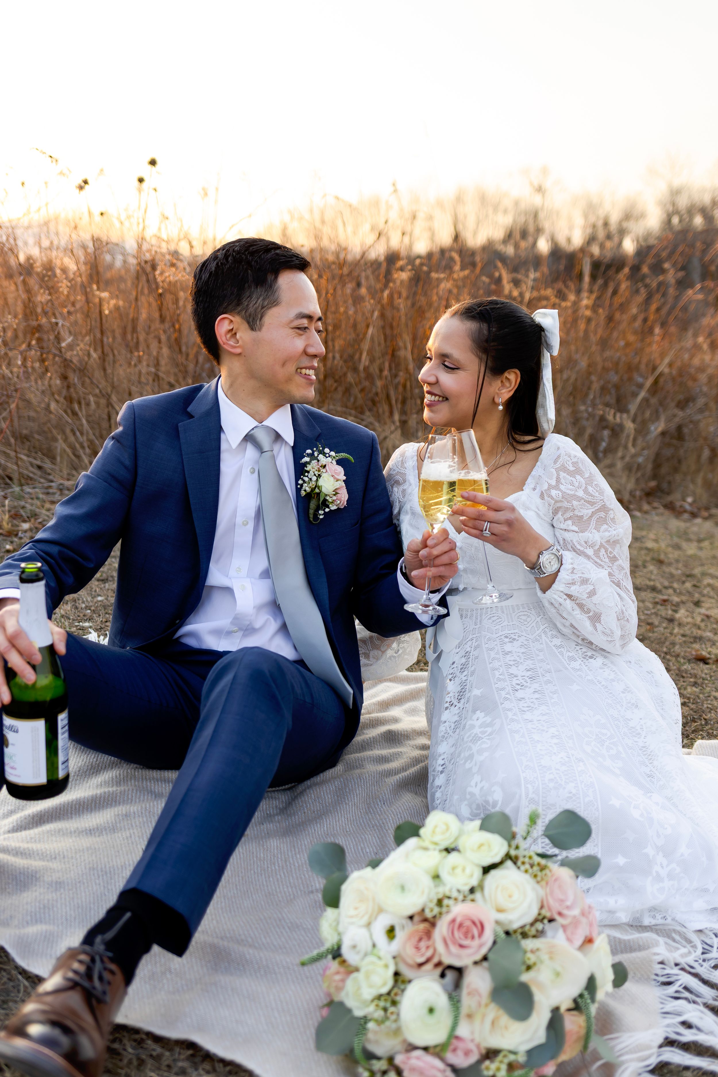 Newlyweds celebrate with champagne outdoors while sitting on blanket with pink and white rose bouquet at sunset.