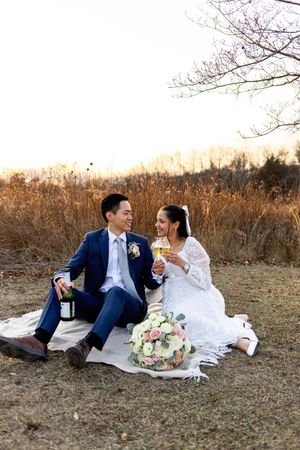 Couple sits on blanket having champagne picnic at sunset in field with bouquet and fall foliage in background.