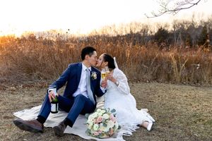 A couple in wedding attire sits on a blanket in a field at sunset with a floral bouquet between them.