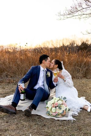 A romantic couple shares champagne during a sunset picnic with a wedding cake and flowers on a blanket.