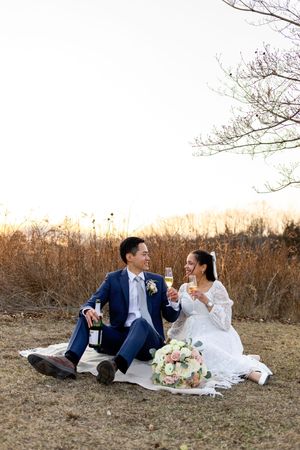 A couple enjoys a romantic picnic in a field at sunset, sitting on a blanket with wine and a floral bouquet.