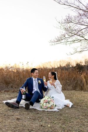 Couple in wedding attire share drinks while sitting on picnic blanket in rustic outdoor setting at sunset.