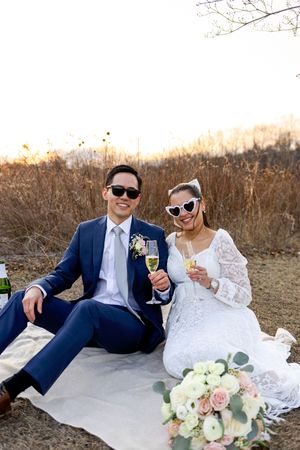 A couple in wedding attire share champagne while sitting on a picnic blanket with a bouquet of roses and peonies nearby.
