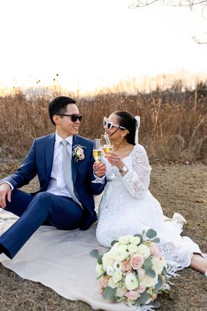Newlywed couple toasting with champagne outdoors on blanket with bridal bouquet of pink and white roses.