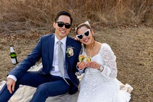 A couple in wedding attire having drinks during an outdoor picnic, wearing sunglasses and smiling together.