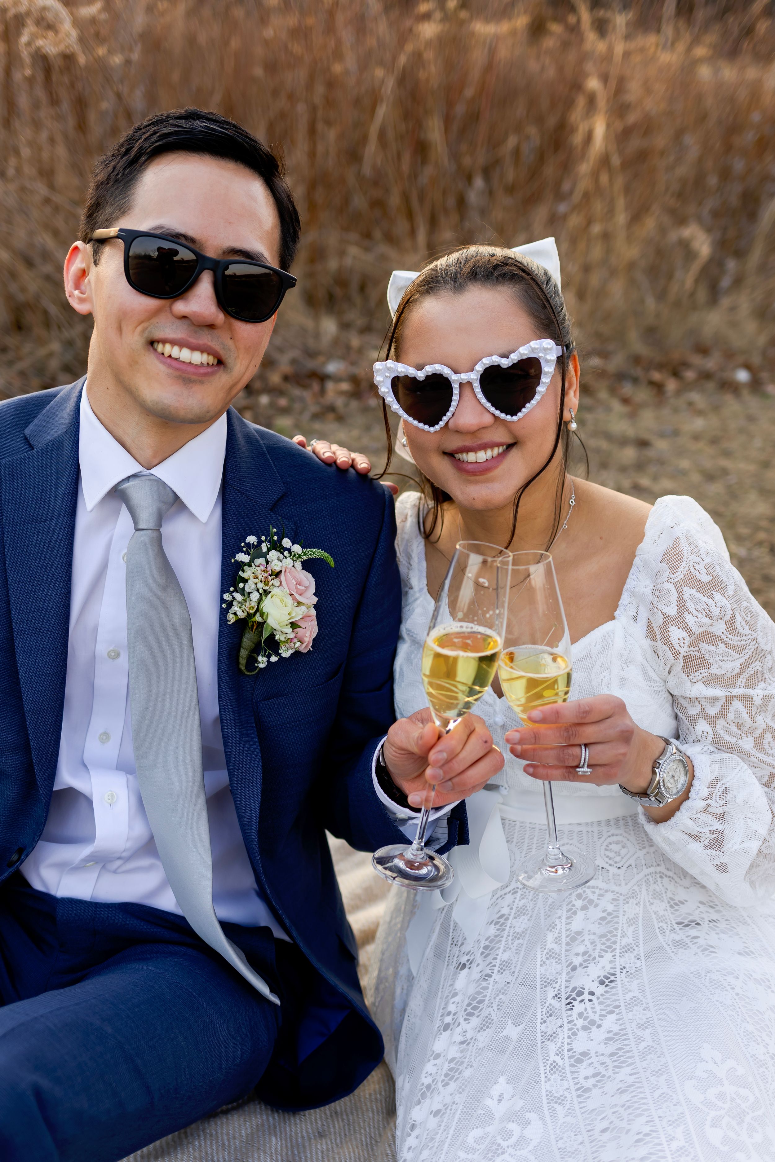 Wedding couple in sunglasses toasting with champagne, wearing navy suit and white lace dress outdoors.
