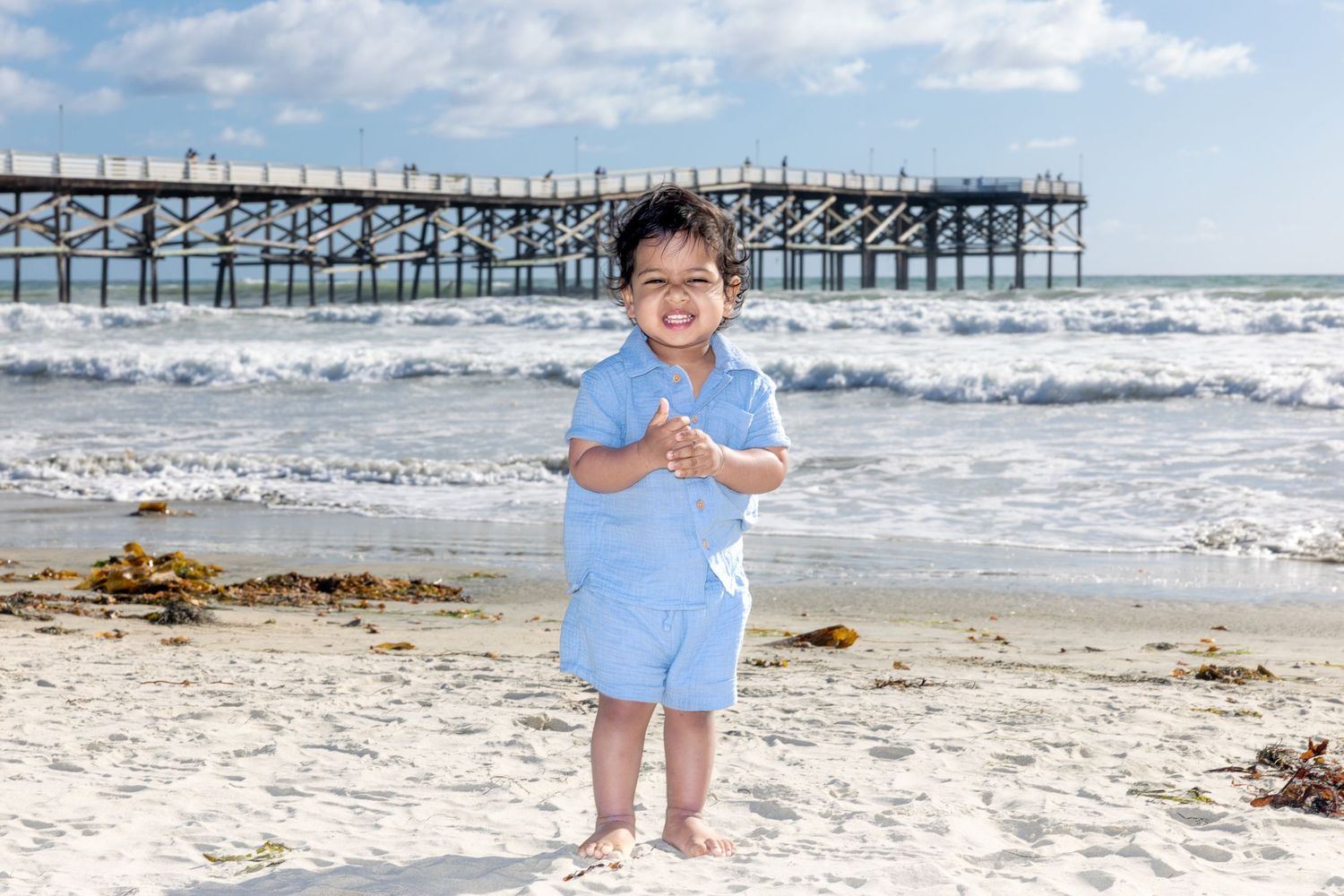 San Diego family photography of a smiling toddler boy standing on the beach with the ocean and wooden pier in the background.