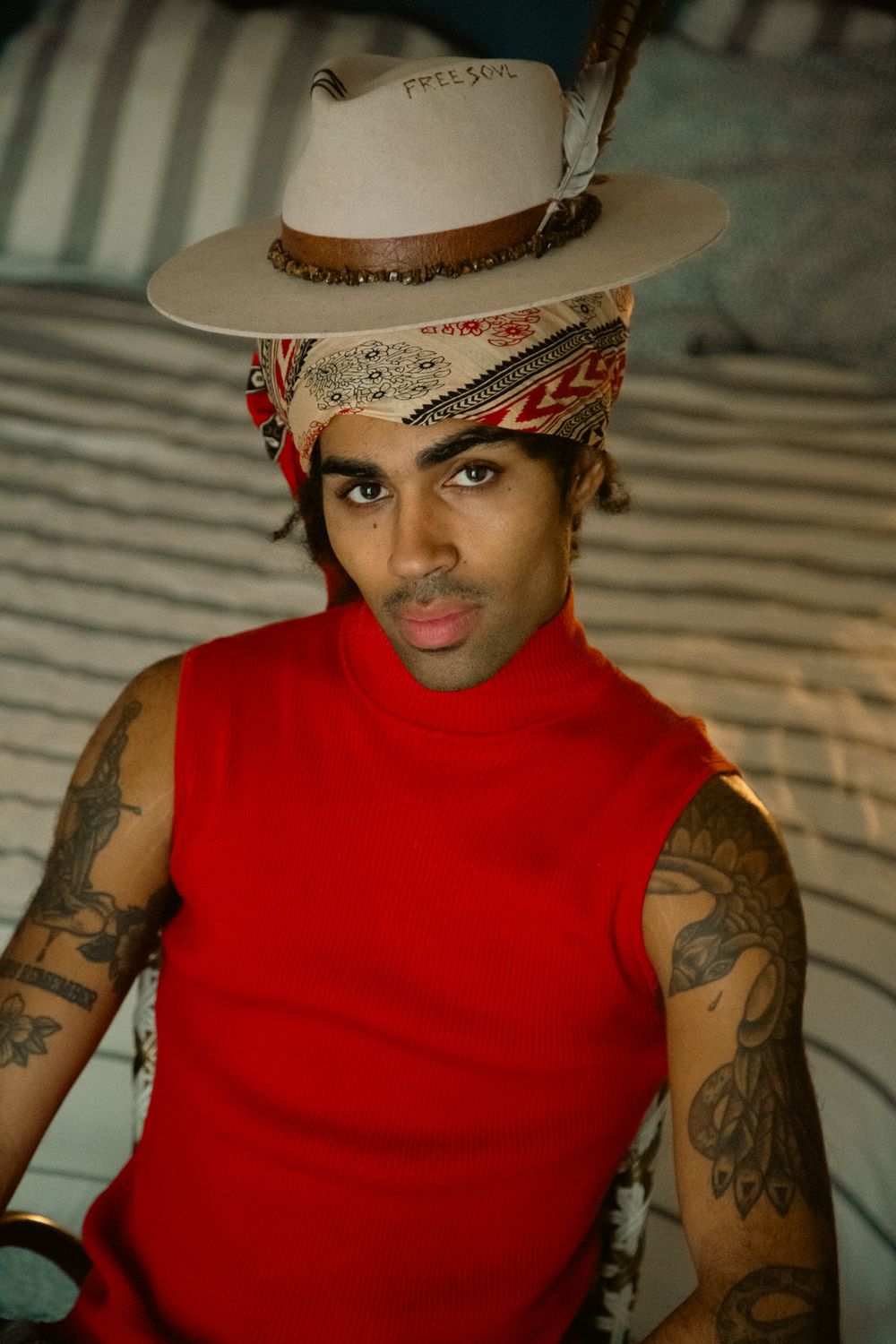 Stylish fashion portrait featuring sleeveless red turtleneck, beige fedora, patterned headscarf and visible arm tattoos.