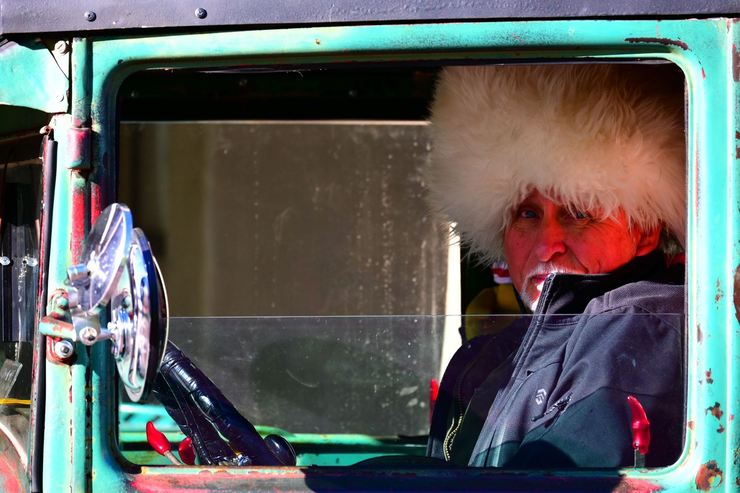 A man with a fur hat peers out from the half-rolled down window of an antique truck.