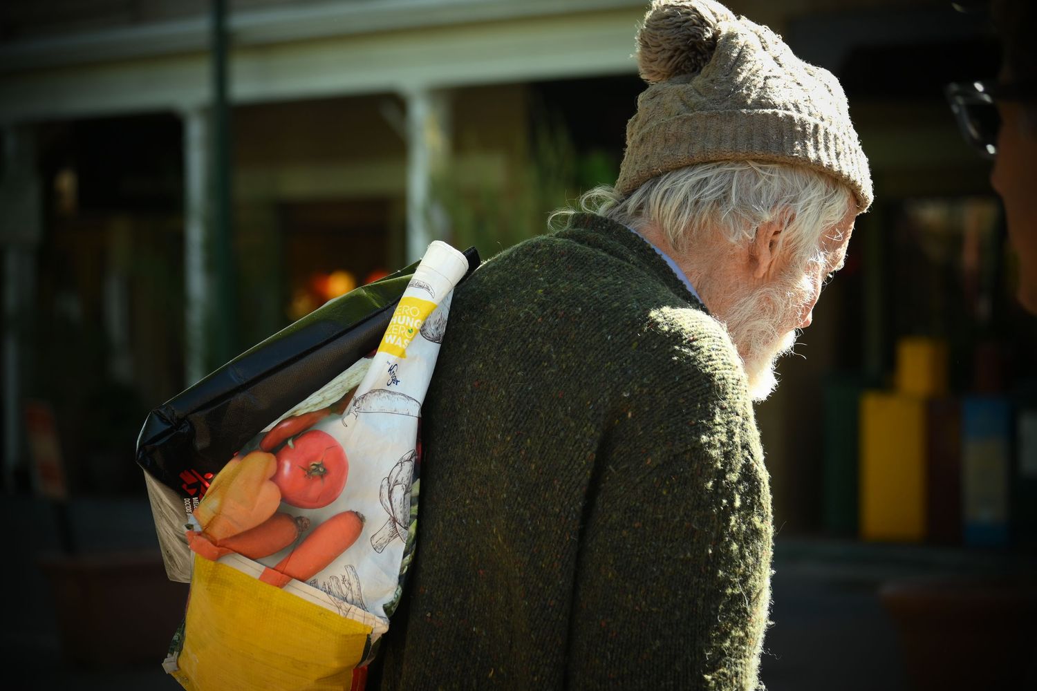 A man wearing a sweater and toboggan hat carries a bag over his shoulder and faces away as he crosses an empty street.