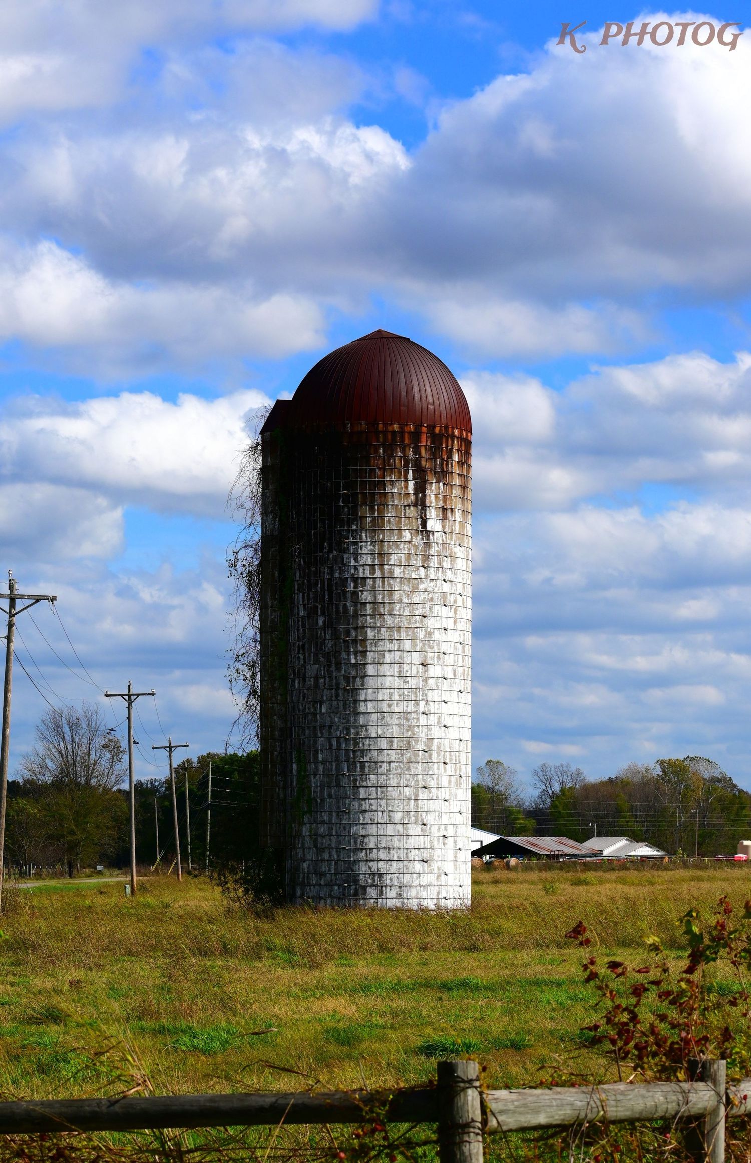 A rusted silo stands tall in a field of yellow grass against a blue sky with white puffy clouds.