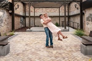 Man lifting woman in a romantic embrace on a brick courtyard outside a rustic venue.