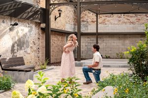 Man proposing on one knee to woman in floral dress in a charming outdoor courtyard with brick walls.