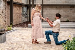 Man kneeling on one knee proposing to woman in floral dress on brick patio outdoors.