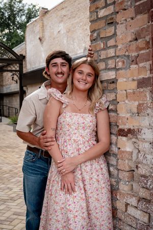 A smiling couple poses against a rustic brick wall, the woman in a floral dress and the man in a beige shirt and jeans.