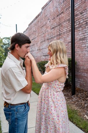Young couple in romantic moment, man kissing woman's hand outdoors near brick wall, woman in floral dress smiling.