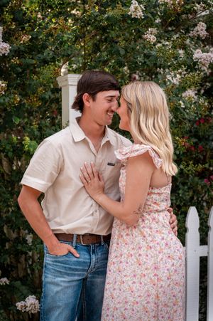 A couple shares a romantic moment in a garden, the woman in a floral dress showing her engagement ring.