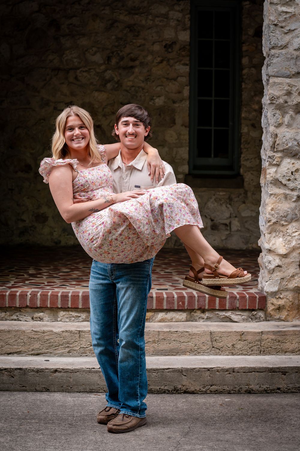 A smiling young man carries a woman in a floral dress on brick steps outside a stone building.