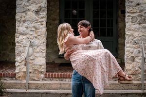 A joyful couple shares a romantic moment as a man lifts a laughing woman in a floral dress outside a stone building.