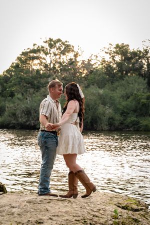 Couple dancing by a river at sunset, woman in white dress and cowboy boots, man in jeans and casual shirt.