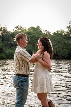 A young couple holds hands and smiles at each other by a river, she in a white dress, he in a striped shirt.