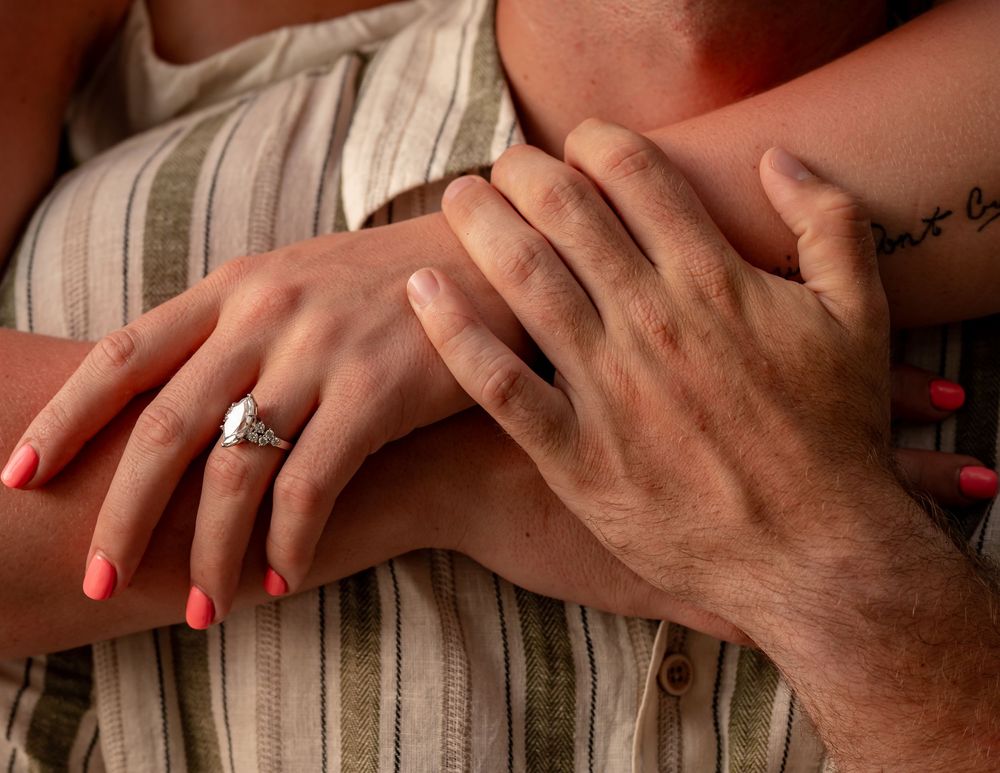 Close-up of couple's hands showing diamond engagement ring with pink nails and tattoo visible on wrist.