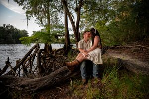 Couple sharing a romantic kiss while sitting on driftwood by a serene river surrounded by lush trees.